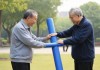 -公园晨练，退休工人张建国在社区公园带领一群老年人进行健身活动，使用自制的大号健身器材锻炼身体，展现老年人积极向上的精神风貌